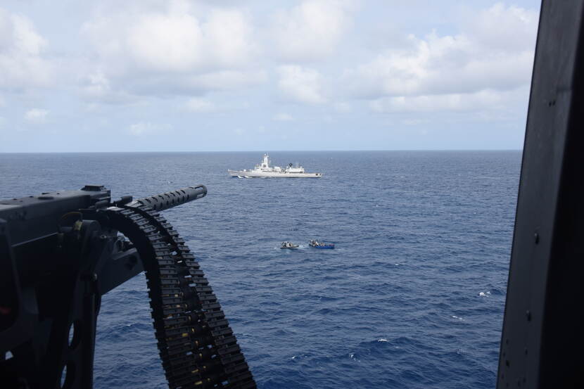 Een foto vanaf een boot met een mitrailleur in beeld. Verderop op zee vaart een andere boot, de mitrailleur lijkt daarop gericht.