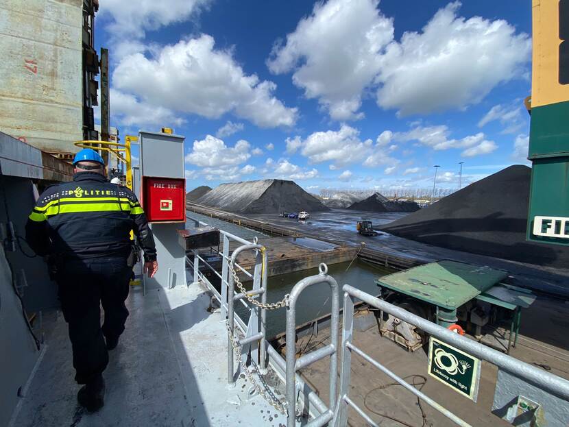 Een politieman is op de rug te zien terwijl hij op hoogte langs de rand van een platform op zee loopt. Over de reling is de kade te zien, daarop liggen donkere bergen grondstof.