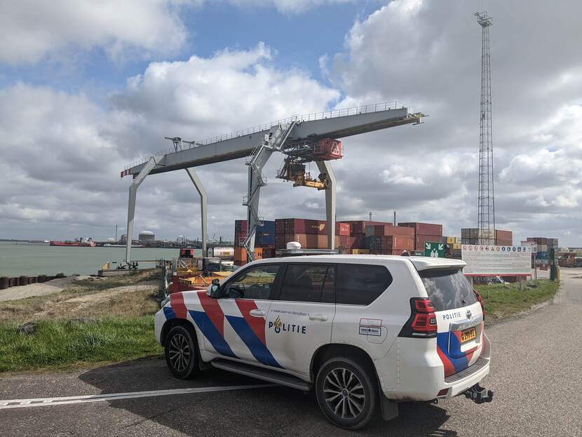 Politieauto staat geparkeerd op de kade. Neus van de auto wijst richting het water en in de achtergrond zijn havencontainers te zien.