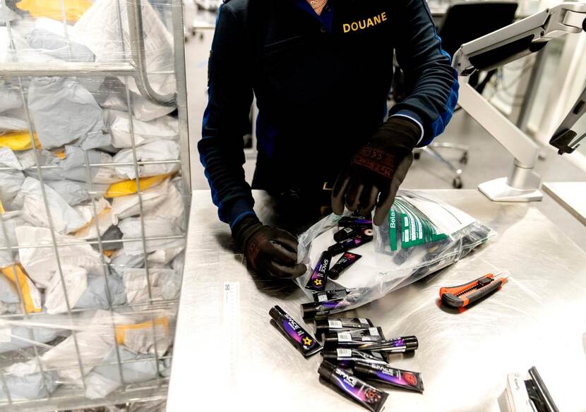 Iemand van de douane opent een plastic die gecontroleerd moet worden. Er komen kleine tubes uitvallen.